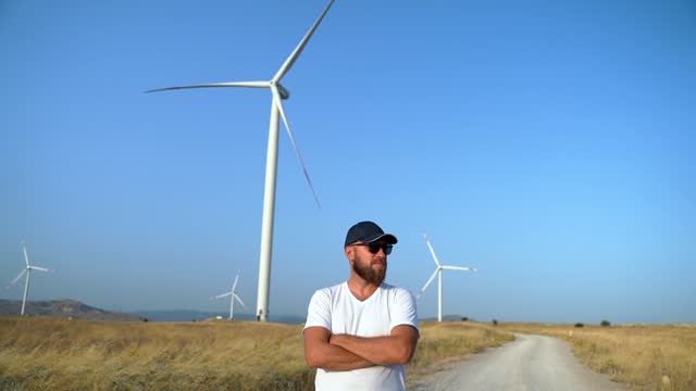 Happy confident man standing cross arms over the chest on country road in the field with windmill wind generator. Freedom, Environmentally eco friendly energy power. sustainable consumption
