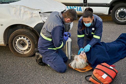 Two uniformed paramedics in protective masks and gloves caring for mature woman lying on spinal board in middle of urban road following crash.