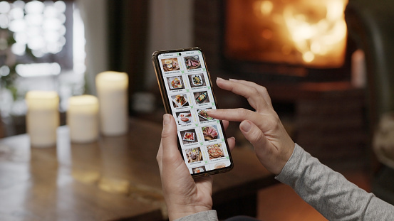 Ordering food using a smartphone at home. A woman selects estaurant food in the internet menu of a gourmet restaraunt using an application on a smartphone. Home evening furnishings with a burning fire in the fireplace.