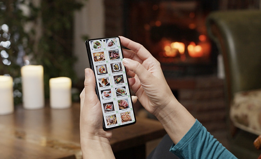 Ordering food using a smartphone at home. A woman selects estaurant food in the internet menu of a gourmet restaraunt using an application on a smartphone. Home evening furnishings with a burning fire in the fireplace.