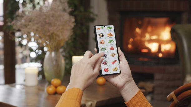 istock Ordering food using a smartphone at home. A woman selects vegetables in an online store using an application on a smartphone. Home evening furnishings with a burning fire in the fireplace. 1572097140