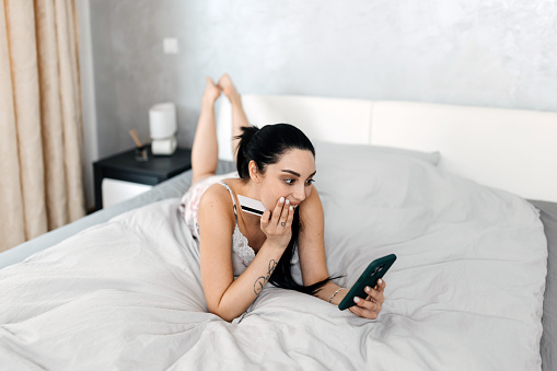 Embracing the convenience of modern technology, the woman lays in bed, utilizing her credit card and mobile phone to browse and purchase online