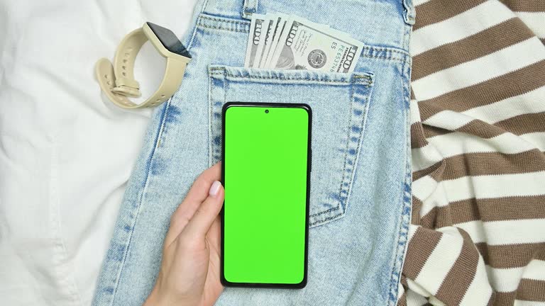 Blue jeans and sweaters with dollars in the pocket, and a smartphone with green screen,female hands tapping on screen, top view.