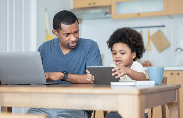 Handsome African American father using laptop computer and his cute little boy using digital tablet learning online, watching video while sitting on table in kitchen room at home. Technology concept.