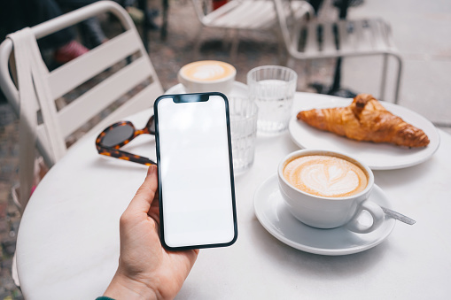 Woman holding smartphone with blank screen in a cafe. Cappuccino and croissant in the background on the table.