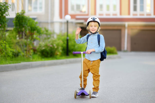 little child in safety helmet riding scooter to school. preschooler boy waving hand saying hi. safety kids by way to school. back to school concept. - little boys preschooler back to school backpack imagens e fotografias de stock