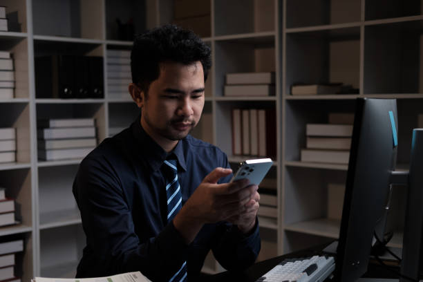 istock Asian businessman using smartphone while working overtime at his office room at night. 1572299878