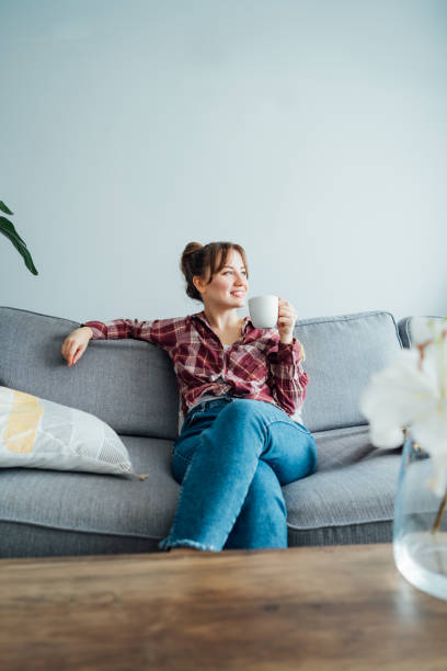 istock Young smiling woman sitting on sofa and looking side up while drinking coffee. Young brunette woman relaxing after housekeeping, home cleaning. Portrait of relaxed female resting at home. Vertical. 1572300955