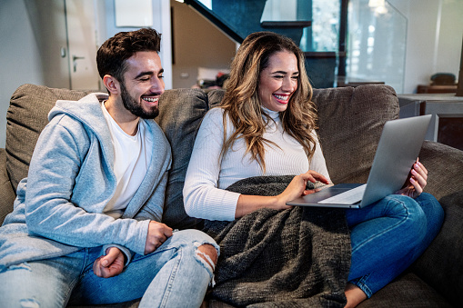 Close-up of a caucasian young couple in their living room watching TV while the wife is using the laptop computer. Lifestyle of a couple on a romantic evening. Domestic lifestyle.