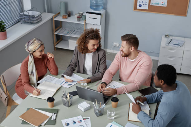 istock Young confident businessman in pink pullover talking to female colleagues 1572376894
