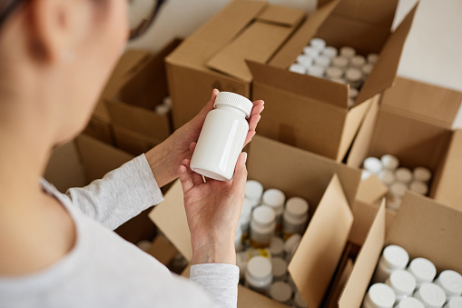 Top view of young woman holding blank pill bottle while packaging medicine at pharmaceutical factory, copy space