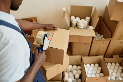 Close up of black young man taping boxes while working in packaging department at factory or shipping services, copy space
