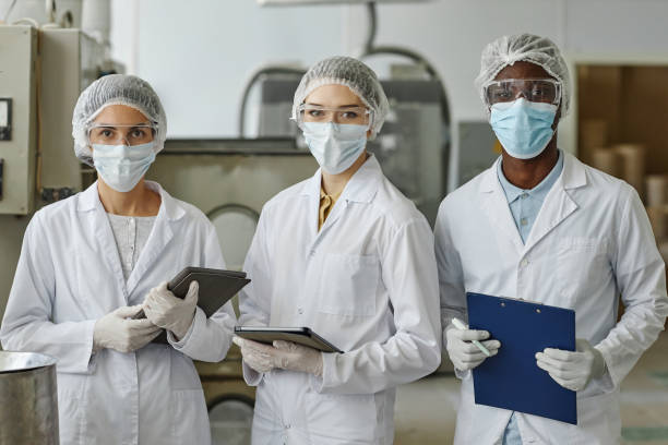 istock Three factory workers wearing lab coats and masks looking at camera in workshop 1572385013