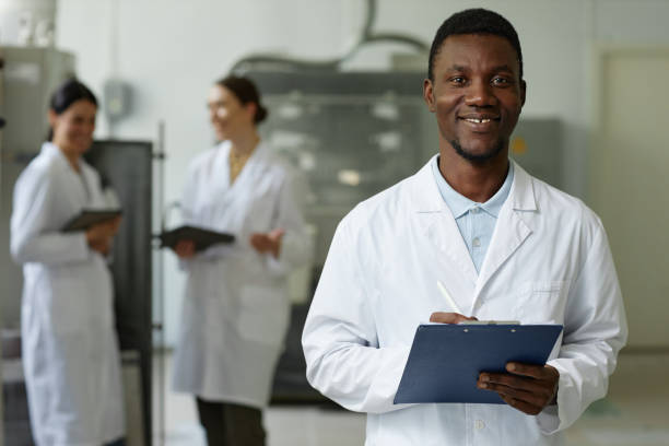 lächelnder schwarzer mann im laborkittel schaut in der fabrikwerkstatt in die kamera - healthcare and medicine smiling group of people lab coat stock-fotos und bilder