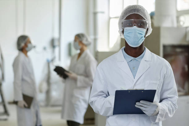 istock Young black man wearing mask and lab coat looking at camera in factory workshop 1572385030