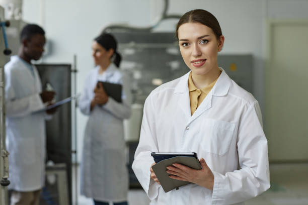 jeune femme souriante en blouse de laboratoire regardant la caméra dans l’atelier de l’usine - manufacturing occupation photos et images de collection