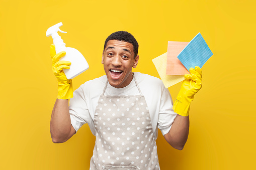 guy african american cleaner in apron and gloves shows rags and detergent on yellow isolated background, cleaning service worker holds sponges and spray
