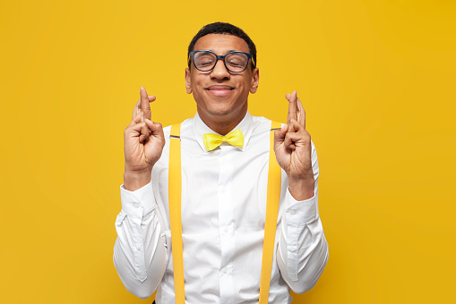 young guy african american in white shirt with suspenders and bow tie hopes and crosses his fingers on yellow isolated background, nerd man waits and hopes for good luck