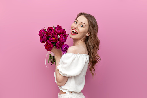young attractive girl in white clothes holds bouquet of pink tulips and smiles, woman on holiday with flowers on pink isolated background, concept of spring and March 8