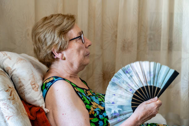 istock Elderly woman sitting sofa home hand fan 1572410320