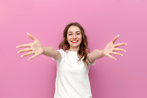 young cute woman in white t-shirt with outstretched arms wants hug, the girl hugs and holds her hands in front of her on pink isolated background