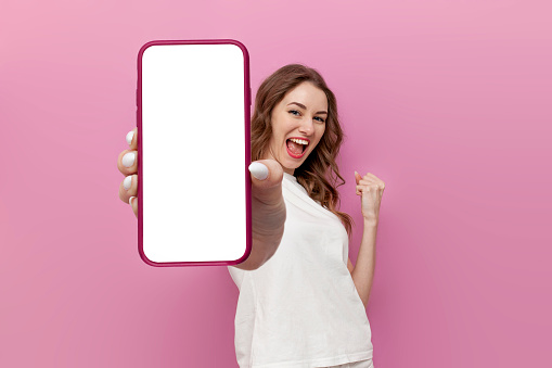young cute woman in white t-shirt shows blank smartphone screen and rejoices in victory on pink isolated background, the winner girl advertises copy space on the phone, mockup