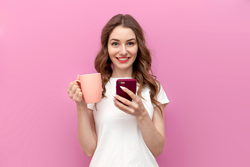 young cute woman in white t-shirt holds cup with drink and uses smartphone on pink isolated background, the girl drinks coffee and types on the phone