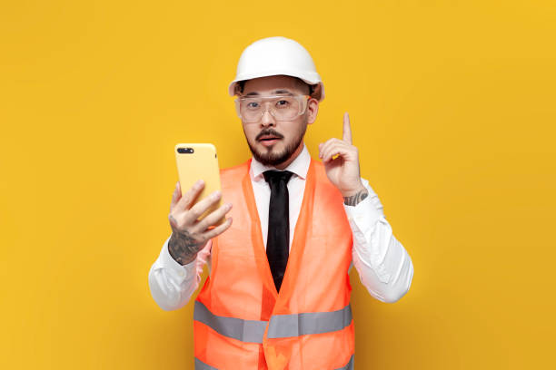 istock asian foreman in uniform holding smartphone and pointing idea with his finger on yellow isolated background 1572419998