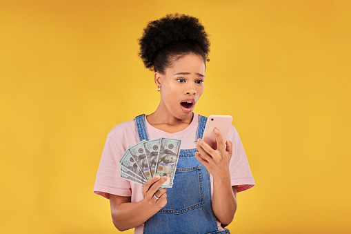 Surprise, money and winner with black woman and phone in studio for success, prize or deal. Bonus, lottery and online payment with person and cash on yellow background for dollar, profit or giveaway