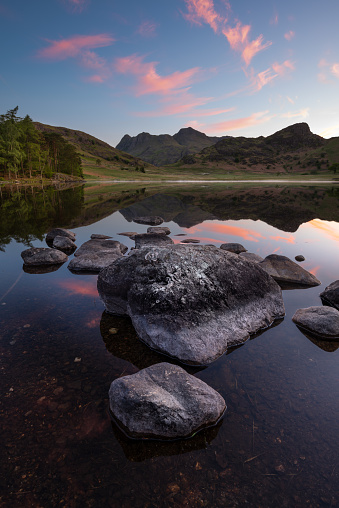 Beautiful pink sunrise clouds high in the sky at sunrise on a Summer morning at Blea Tarn in The Lake District, UK. Mirror like reflections can be seen in the water with a breathtaking view of The Langdale Pikes in the background.