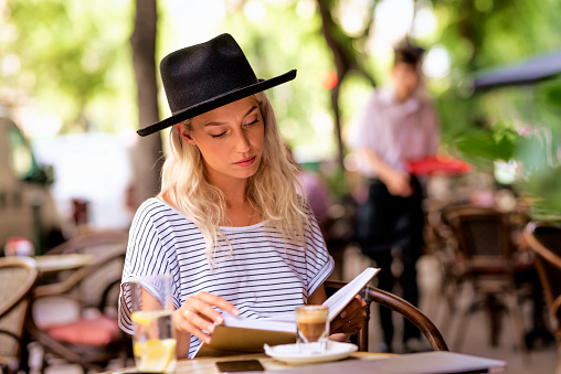 A woman with blonde hair and a hat is sitting on the terrace of a cafÃ© and reading a book.