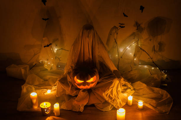 istock Spooky ghost holding Jack o lantern on background of spider web with bats and glowing lights in dark. Scary atmospheric halloween party decorations and person under sheets. Happy Halloween! 1572478384