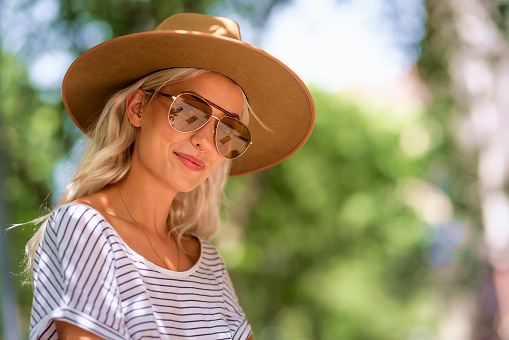 A young girl with blonde hair wearing sunglasses and a hat sitting on a cafÃ© terrace. Attractive female looking at camera and smiling.