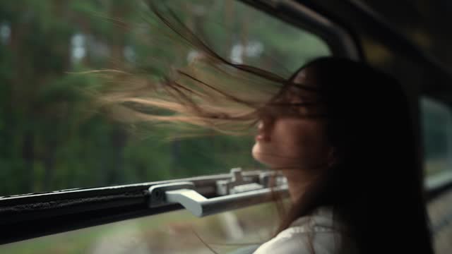 Close up view of  young long-haired woman or girl looks out of an open train window while the train is moving. Hair blowing in the wind. Enjoys traveling by train, a trip to the mountains in the summer.