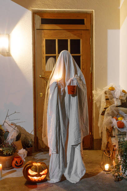 istock Scary ghost with halloween candy bucket and glowing Jack o lantern at doors in spooky night. Happy Halloween! Person dressed as ghost trick or treating on porch with halloween decorations 1572481986