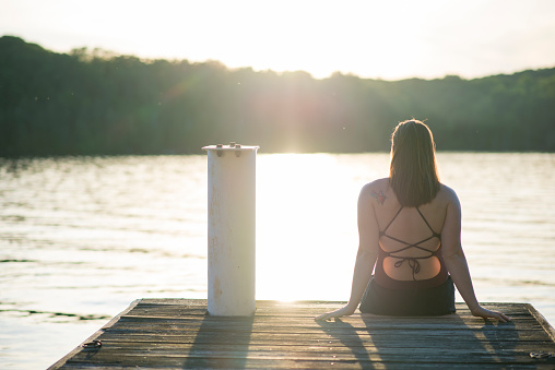 Relaxed anonymous young woman with brown hair is stretching out in a bathing suit and looking out at the lake water gently at sunset. She is sitting on a wooden dock at a cottage. The woman has a small tattoo on her shoulder.