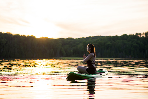 Side profile of a beautiful young woman in a modest bathing suit sitting on a stand up paddleboard floating in the middle of the lake at sunset in the summer. She is sitting cross-legged and doing prayer position in yoga as she is finding meditation and relaxation.