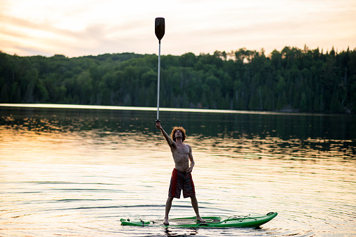 Young man with long curly hair is holding up a paddle with the tips of his fingers while balancing on a stand up paddleboard at sunset in the summer at the cottage on a lake.