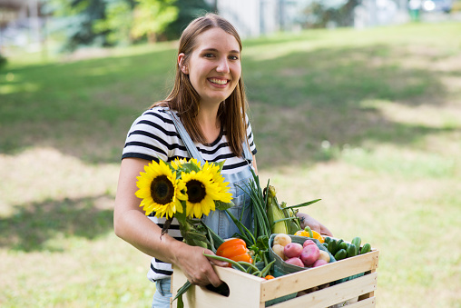 Young female farmer wearing a striped shirt and jean bib overalls is standing outdoors at a local farmers market on the grass holding a wooden crate full of freshly harvested organic flowers, fruits and vegetables from the farm,