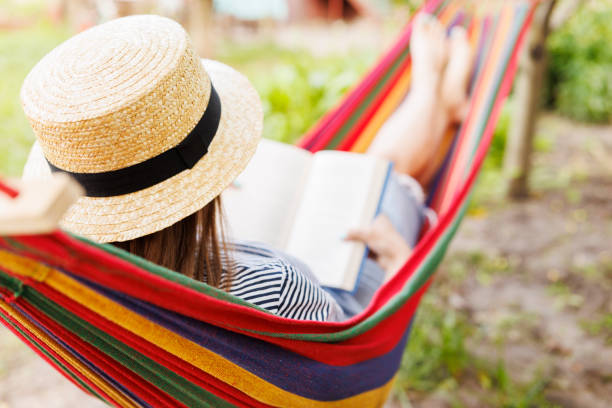 istock Young woman reading book while lying in comfortable hammock at green garden 1572539730