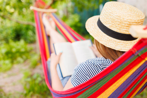 istock Young woman reading book while lying in comfortable hammock at green garden 1572540248