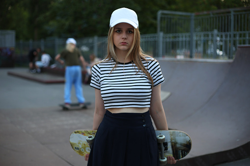 Young European blonde student girl in a topic and a white cap holds a skateboard on the background of a skateboard with jumping ramps