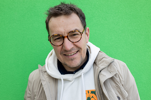 A portrait of a mature man with glasses standing in front of a green wall