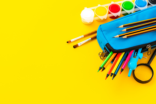 School supplies: pencil case full of colored pencils, compass, paint and magnifying glass on yellow background. Copy space