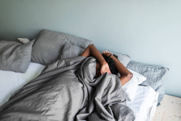 두통을 앓고 있는 우울한 젊은 흑인 여성이 침대에 누워 담요 아래에 숨어 있다 스톡 사진