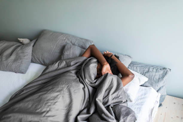 Depressed young adult black woman with headache lying in bed and hiding under blanket Depressed young adult black woman with headache lying in bed and hiding under blanket trouble sleeping stock pictures, royalty-free photos & images