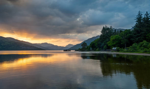 Peaceful twilight scene over a calm lake with smooth water and dark mountain silhouettes on the horizon. Soft gradient sky transitions from warm orange glow near the horizon to deep blue and purple tones, creating a tranquil and minimalist landscape atmosphere.
