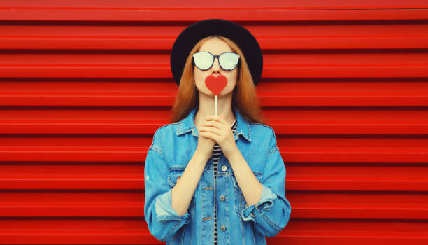 Portrait of beautiful young woman with lollipop blowing her lips with lipstick sending sweet air kiss wearing sunglasses, black round hat on red background stock photo