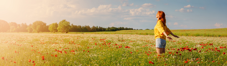 Woman in profile standing on the field with a lot of wild flowers. Panoramic view of the sunset in summer park.