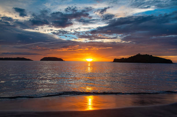 Wide angle landscape photo of a sunset on a beach in Costa Rica. Photo taken on Playa Potrero. The sky is overcast, small islands are visible in the distance as silhouettes and the reflection of the sun is visible in the water and on the wet sandy beach. Beautiful and relaxing.